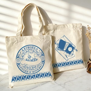 May include: Two off-white canvas tote bags featuring blue ink illustrations. The front bag displays a circular emblem with a creature and the text ARTWORKS ATELIER OF WITCH HAT. The second bag shows a stack of books with a geometric border pattern.