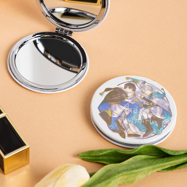 May include: A white, round, compact mirror with a silver rim, open to reveal a reflective surface. A second closed compact mirror with an illustration of three figures and the text "ATELIER OF WITCH HAT" is also visible.