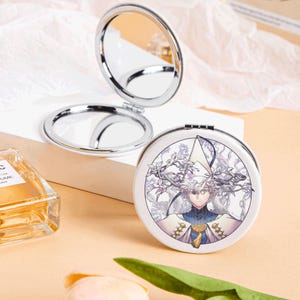 May include: A silver-framed, round compact mirror, open to reveal two reflective surfaces. The closed side features a white background with a detailed illustration of a character in a white and blue outfit. The compact is on a white surface.