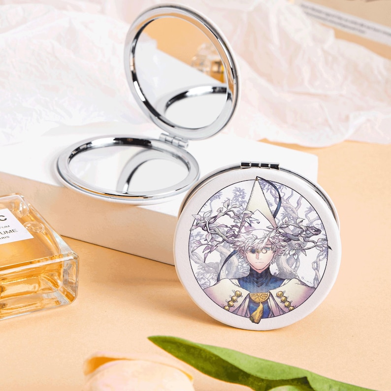 May include: A silver-framed, round compact mirror, open to reveal two reflective surfaces. The closed side features a white background with a detailed illustration of a character in a white and blue outfit. The compact is on a white surface.