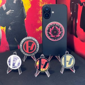 Op de afbeelding: Een zwarte smartphone met een cirkelvormige popsocket met een roze en zwart ontwerp. Vier ronde pins met een gestileerd "Z"-logo in verschillende kleuren worden op transparante standaards getoond. De achtergrond is rood en zwart.