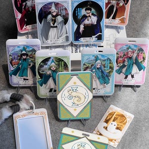 Può includere: Una collezione di porta badge rettangolari in plastica con illustrazioni in stile anime di Witch Hat Atelier, che includono varie figure con cappelli a punta e mantelli, insieme a due supporti che mostrano una piccola creatura bianca.