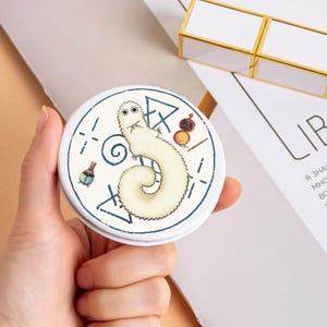 May include: A round, white compact mirror with a cartoon worm design. The worm is cream-coloured with a fluffy body, surrounded by blue geometric shapes and small illustrations. The mirror is held in a hand.