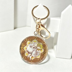 以下が含まれることがあります： ゴールドトーンの金属製リングと留め具が付いた円形のキーホルダー。ペンダントはファセット加工された透明なアクリルディスクで、茶色と黄色の花柄を背景に、金髪の人物が丸まって眠っているイラストが描かれています。