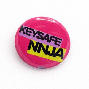 May include: A round, bright pink button with the words "KEYSAFE NINJA" in black, purple, and yellow. The button has a glossy finish and is likely made of plastic. The text is stacked on top of each other.