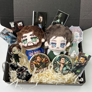 May include: A collection of The Boys merchandise in a black box, including two plush dolls with embroidered faces, two circular coasters featuring Soldier Boy and The Deep, acrylic character stands, and rectangular cards with character portraits.