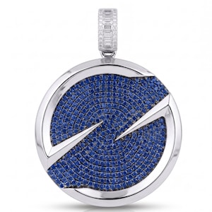 May include: A circular silver pendant featuring a central field of pavé-set blue stones arranged in a swirling pattern. The pendant is framed by a polished silver border with two sharp, pointed silver accents and includes a rectangular bail.