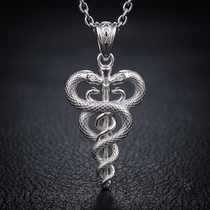 Puede incluir: Un collar con colgante de tono plateado que presenta un símbolo de caduceo con dos serpientes entrelazadas alrededor de un bastón central. Las serpientes tienen escamas texturizadas y pequeñas piedras claras en sus cabezas. El colgante cuelga de una cadena.