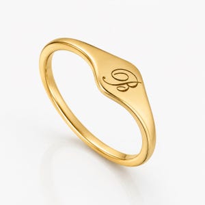 May include: A gold signet ring with a polished finish. The ring has a flat, oval face with an engraved script initial "B". The band is smooth and rounded. A classic piece of jewellery.