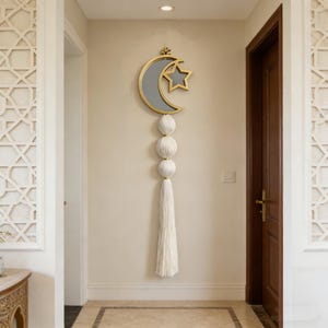 Könnte beinhalten: Wandbehang mit goldfarbenem Halbmond- und Stern-Design, grauer Innenraum. Unter Mond und Stern befinden sich drei weiße, kugelförmige Ornamente und eine lange, weiße Quaste. Der Wandbehang hängt an einer beigen Wand.