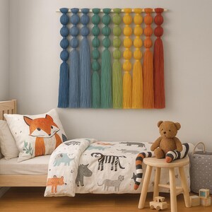 May include: A children's bedroom scene featuring a colorful wall hanging, a bed with a fox pillow and animal-themed bedding, a teddy bear, and a small wooden stool. The wall hanging has blue, green, yellow, and orange tassels.