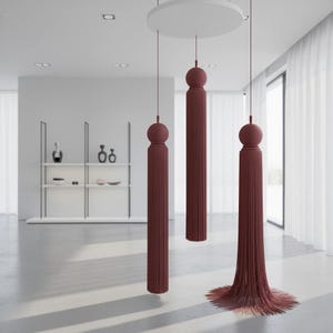 May include: Three hanging decorative tassels in a deep maroon colour, each featuring a spherical top and long, straight, fine fringe. The tassels are suspended from a circular white ceiling mount at varying heights, with the lowest one splayed.