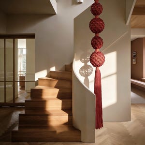 Puede incluir: Una escalera de madera con una pared blanca curva y un adorno colgante rojo decorativo. El adorno tiene cuatro esferas texturizadas y una larga borla. Las escaleras son de madera clara, y el suelo tiene un patrón de espiga.