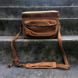 May include: A brown suede camera bag with a long adjustable strap. The bag has a zippered pocket on the front and a flap closure.