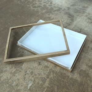 May include: An open, square display box with a clear glass top and a light-colored wooden frame. The box is white inside and sits on a concrete surface. The glass top is slightly ajar, revealing the interior.
