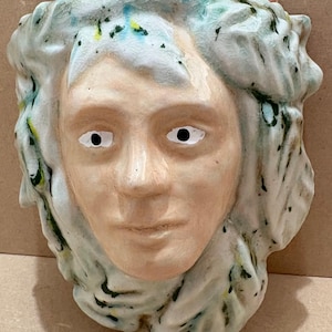 May include: A ceramic face sculpture with a calm expression. The face is a pale peach colour with white eyes and black pupils. The hair is a textured, light blue-green with darker green and yellow accents. Suitable for home decor.