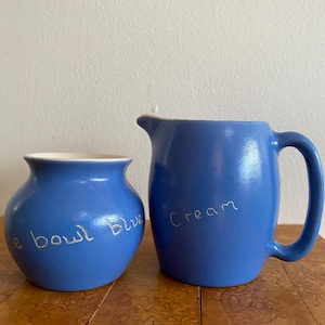 May include: Two blue ceramic pieces: a small bowl with "e bowl blue" and a pitcher with "Cream" in handwritten script. The pitcher has a handle and spout. Both pieces have a matte finish and a white interior.