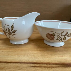 May include: A white creamer and sugar bowl set. The creamer has a handle and spout, while the bowl has a wide rim. Both pieces are decorated with brown floral and leaf patterns. The interior of the creamer and the rim of the bowl are a light pink color.