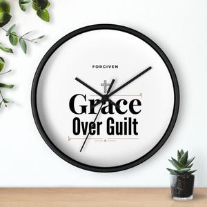 May include: A round wall clock with a black frame and a white face. The clock features the words "Grace Over Guilt" in a bold, black font, with a cross symbol above. The word "Forgiven" is above the cross.