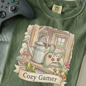 May include: Green t-shirt with a graphic of a watering can, a video game controller, and the words "Cozy Gamer" on a banner. A gray video game controller is in the upper left corner. The shirt has a "Comfort Colors" tag.