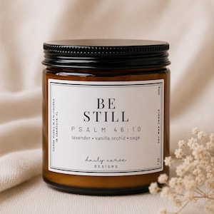May include: A soy wax candle in a brown glass jar with a black lid. The label reads "BE STILL" with "PSALM 46:10" and scent notes. The candle is 7 oz and hand-poured in Sarasota, FL.