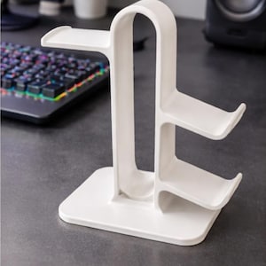 May include: A white headphone stand with a rectangular base and curved supports. The stand has two angled shelves for holding headphones and a top platform. The stand is designed to sit on a desk or table.