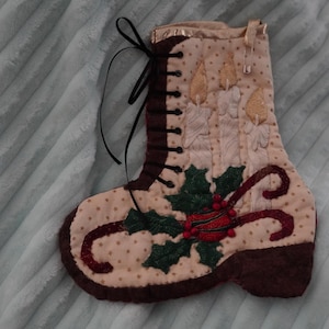 Peut inclure: Une chaussette de Noël en tissu matelassé en forme de botte, avec une base crème à motif d'étoiles, une bordure marron foncé, un ruban noir à lacets et une broderie représentant trois bougies allumées, du houx vert et des baies rouges.