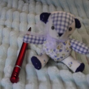 Puede incluir: Un pequeño oso de peluche con un patrón de cuadros blancos y azules en la cabeza y los brazos, un estampado floral en el torso y detalles en azul oscuro en las orejas y los pies. Un bolígrafo rojo está junto al oso como referencia de escala.