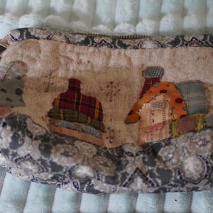 Pode incluir: Uma bolsa com fecho feita de tecido bege com textura acolchoada e aplicações decorativas de tecido de pequenas casas em padrões xadrez e de bolinhas. A bolsa é rematada com um tecido de padrão floral cinzento escuro e branco nas bordas.