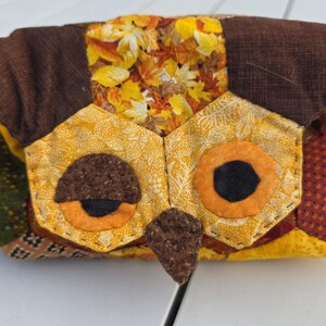 May include: A fabric pouch shaped like an owl, featuring a patchwork design in shades of brown, orange, yellow, and green. The owl face has two large yellow eyes with black pupils, a dark brown beak, and a forehead patterned with autumn leaves.