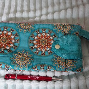 May include: A rectangular teal fabric pouch featuring a repeating orange, white, and brown mandala pattern, a side wrist strap, and a teal flap secured with a gold snap button. A red pen with the white text AGEDI is positioned below the pouch.