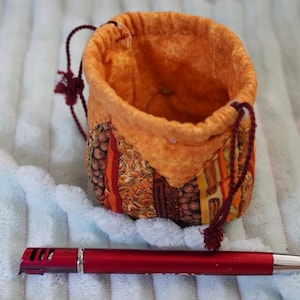 May include: A small fabric drawstring pouch featuring a vibrant orange, yellow, and brown patterned exterior with a solid orange interior lining. The pouch is positioned next to a red ballpoint pen for scale, showing its compact size.