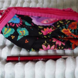 Può includere: Una pochette in tessuto nero con un vivace motivo floreale nei toni del rosa, arancione, turchese e giallo, rifinita con una cerniera rosa brillante. Una sottile penna in metallo rosso è posizionata sotto per riferimento di scala.