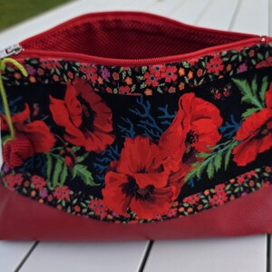 Può includere: Una pochette con cerniera caratterizzata da un pannello in tessuto nero con una vivace stampa floreale di papaveri rossi, accentuata da un bordo floreale più piccolo. La parte inferiore è in materiale rosso tinta unita e l'interno è foderato in rete rossa.