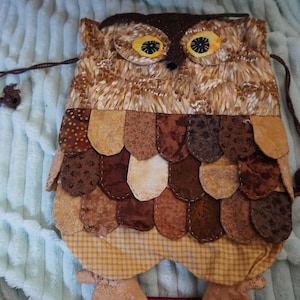 May include: A drawstring pouch shaped like an owl, featuring a textured brown and tan fabric body with layered, scalloped fabric pieces in various shades of brown and beige to mimic feathers. It has yellow eyes with black pupils and brown feet.