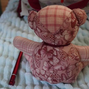 Op de afbeelding: Een knuffelbeer gemaakt van roze stof met een vlinderpatroon en een ruitjesontwerp op het hoofd en de armen. Een dun donker lint zit om de nek. Een rode pen ligt naast de beer om een referentie te geven voor de schaal.