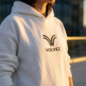 May include: A white hoodie with the word "VOLVEZ" printed in black. The hoodie has a drawstring hood and ribbed cuffs. The logo is a stylized V-shape.