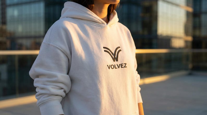 May include: A white hoodie with the word "VOLVEZ" printed in black. The hoodie has a drawstring hood and ribbed cuffs. The logo is a stylized V-shape.