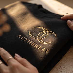 Könnte beinhalten: Schwarzes T-Shirt mit goldener Stickerei, die zwei ineinandergreifende Halbmonde und das Wort "AETHERIAL" zeigt. Das Shirt ist gefaltet und wird von Händen gehalten. Das Design ist auf der Brust zentriert.