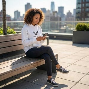 May include: A person wearing a white hoodie with the word "VOLVEZ" in black, sitting on a wooden bench. The person is wearing black jeans and gray sneakers, holding a smartphone. The background features a cityscape and a sunny day.