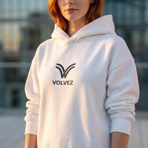 May include: A white hoodie with the word "VOLVEZ" printed in black. The hoodie has a hood and long sleeves. The logo is a stylized V-shape. The person is standing in front of a building.