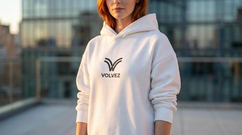 May include: A white hoodie with the word "VOLVEZ" printed in black. The hoodie has a hood and long sleeves. The logo is a stylized V-shape. The person is standing in front of a building.