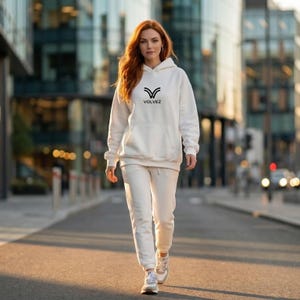 May include: A person wearing a white hoodie and matching sweatpants. The hoodie has a black logo and the word "VOLVEZ" printed on the front. The person is walking on a paved road with buildings in the background.
