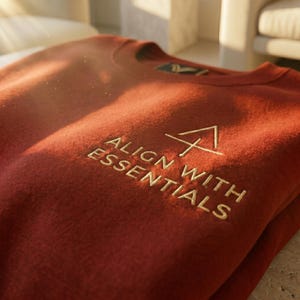 May include: A rust-colored sweatshirt with the embroidered text "ALIGN WITH ESSENTIALS" and a geometric design. The crew neck sweater is folded, with the text and design in a lighter shade of gold.
