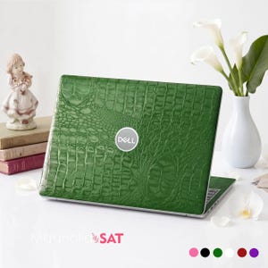 Könnte beinhalten: Ein Laptop mit einer grünen Abdeckung in Krokodilleder-Optik und einem kreisförmigen silbernen Dell-Logo in der Mitte. Das Gerät steht auf einer weißen Oberfläche neben einer kleinen Figur, gestapelten Büchern und einer weißen Blumenvase.