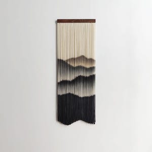 May include: A rectangular wall hanging with a wooden top and long, vertical strands. The strands transition from cream to brown and black, creating a mountain-like design. The bottom edge is a solid black.