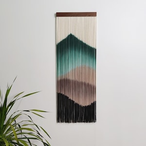 Könnte beinhalten: Ein Wandbehang mit einer dunkelbraunen Holzleiste an der Oberseite und langen, vertikalen Baumwollschnüren in einem Farbverlauf. Die Farben wechseln von Cremeweiß oben zu tiefem Waldgrün, gedämpftem Mauve und schließlich Schwarz am unteren Rand.