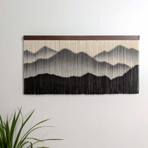 May include: A wall hanging featuring a horizontal dark wood bar from which numerous vertical off-white and black strings are suspended. The strings are arranged to create a layered mountain landscape design with gradients of black, gray, and white.