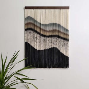 May include: A wall hanging featuring vertical cream-colored cords suspended from a dark wooden bar. The cords are dyed in horizontal, wavy layers of gray, brown, and black, with a speckled texture appearing in the middle section.