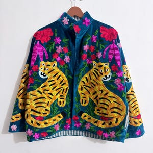 Op de afbeelding: Teal fluwelen jas met geborduurde tijgers, bloemen en bladeren. De jas heeft een mandarijnkraag en een gestreepte voering aan de zoom. De tijgers zijn geel en zwart en de bloemen zijn roze en rood.